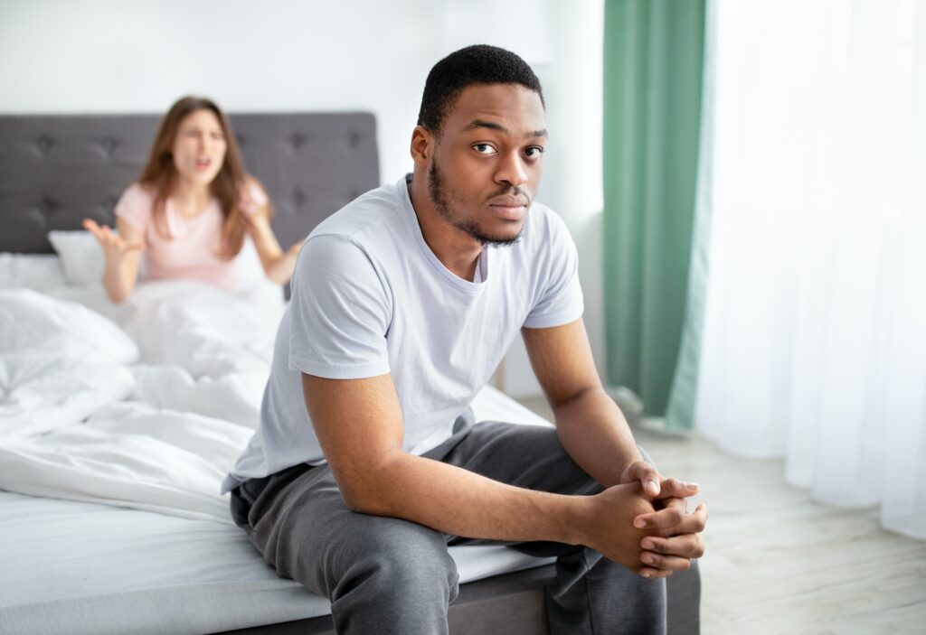 Unhappy black man sitting on bed, overwhelmed with family problems, his angry girlfriend quarelling